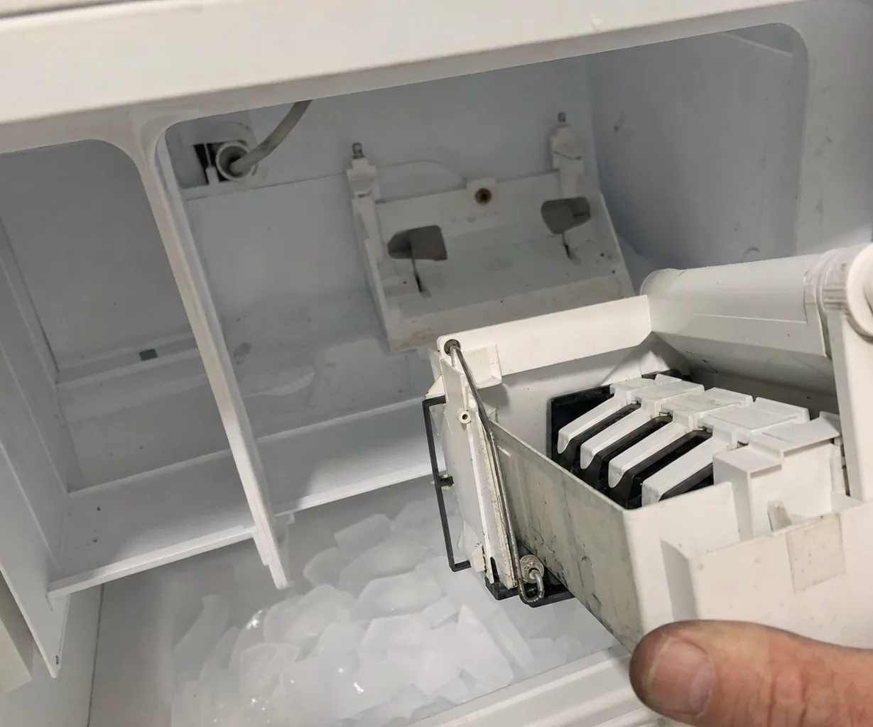 A white ice maker module lifted off its mounting bracket inside a freezer compartment, held by a thumb above a bin of loose cubes, the fill tube fitting visible behind it.