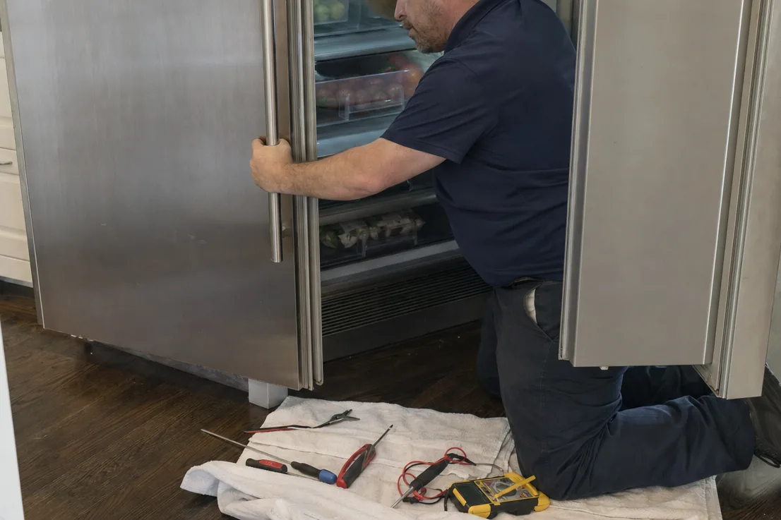 A technician reaching into the lower panel of an open built-in refrigerator, where the control wiring runs