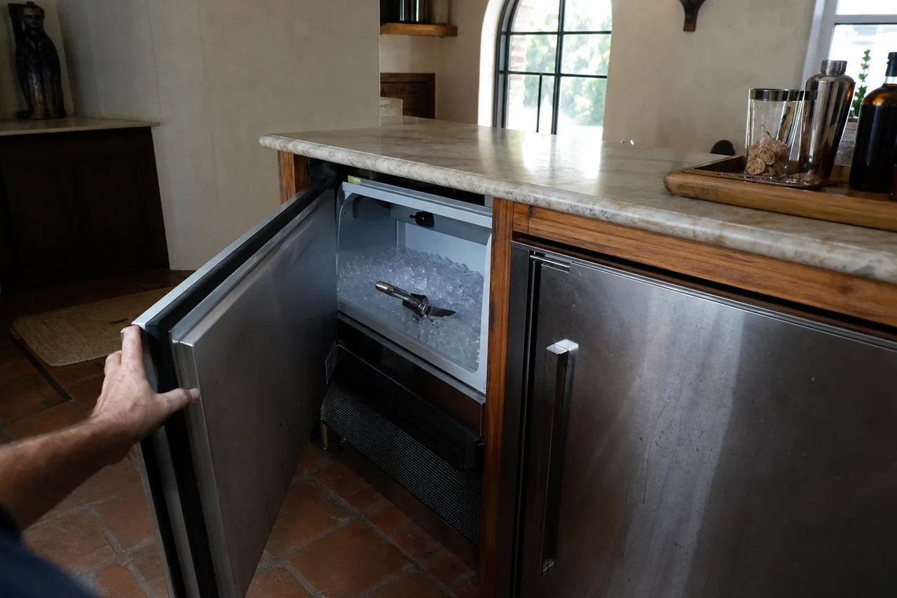 A hand swings open the stainless door of an undercounter ice maker beneath a stone bar top, showing a bin of clear ice with a metal scoop, above terracotta floor tiles